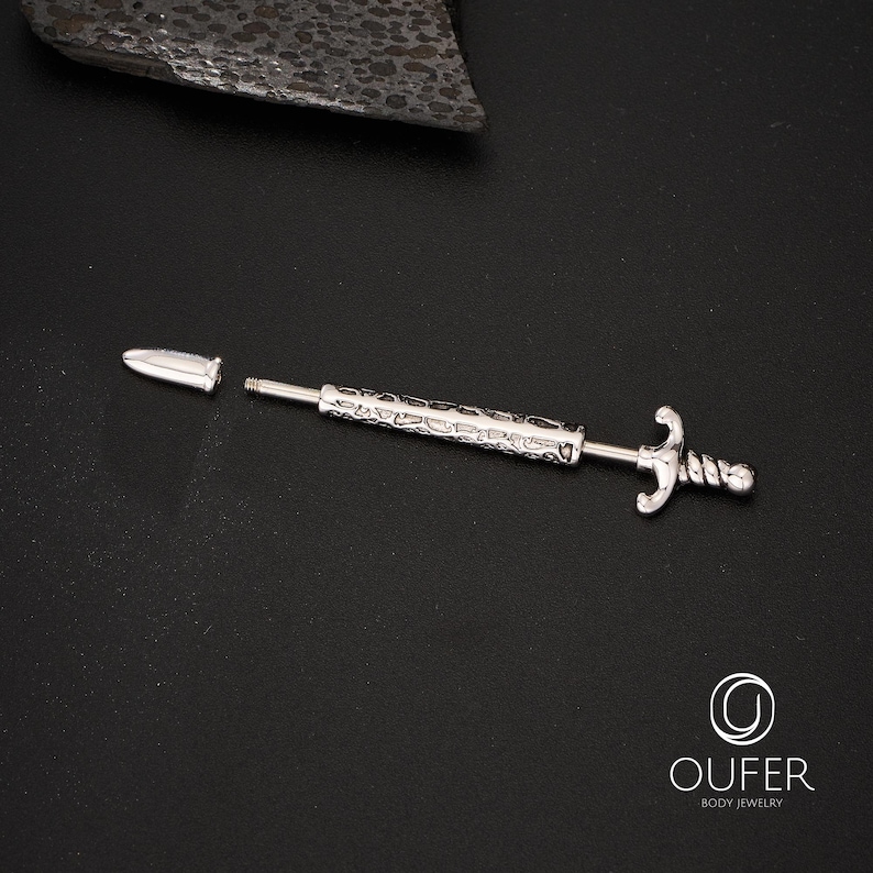 K&ouml;nnte beinhalten: Silberfarbener K&ouml;rperschmuck in Form eines Schwertes, mit strukturiertem Griff und dekorativem Knauf. Der Schmuck ist zerlegt, mit abgetrennter Spitze. Der Hintergrund ist schwarz.