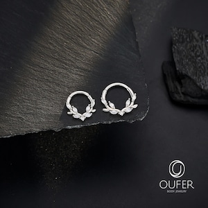 Op de afbeelding: Twee zilverkleurige oorringen met blad- en edelsteenaccenten. De oorbellen worden gepresenteerd op een donkere, getextureerde ondergrond. De oorbellen zijn van Oufer Body Jewelry.