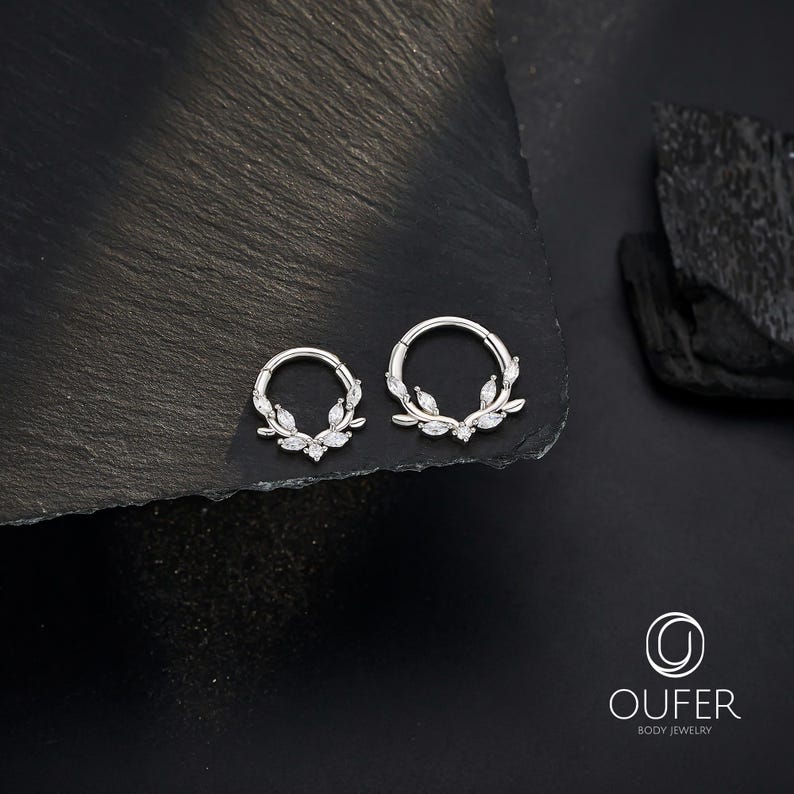 Op de afbeelding: Twee zilverkleurige oorringen met blad- en edelsteenaccenten. De oorbellen worden gepresenteerd op een donkere, getextureerde ondergrond. De oorbellen zijn van Oufer Body Jewelry.