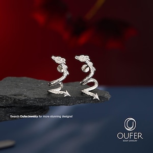 Puede incluir: Un par de pendientes de metal plateado con forma de dragones enroscados con escamas texturizadas y colas puntiagudas. Los pendientes tienen cierre de presión y se muestran sobre una piedra oscura y rugosa. Texto: Search OuferJewelry for more stunning designs! OUFER BODY JEWELRY.
