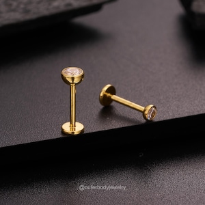 May include: Two gold lip rings with clear gemstones. The rings are on a black surface.