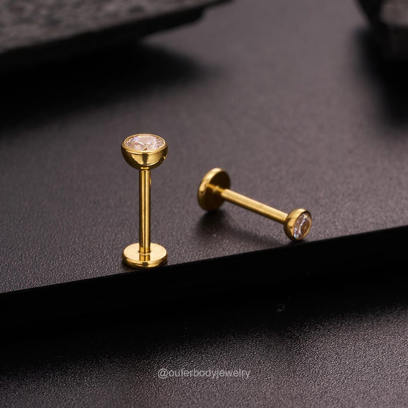 May include: Two gold lip rings with clear gemstones. The rings are on a black surface.