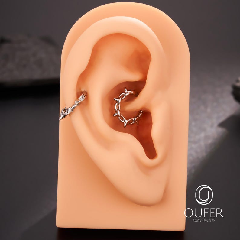 Op de afbeelding: Een zilverkleurige oor piercing sieradenset. E&eacute;n stuk is een ketting en de andere is een hoepel met kleine spikes. De sieraden worden getoond op een huidkleurig oormodel. De merknaam "OUFER BODY JEWELRY" is zichtbaar.