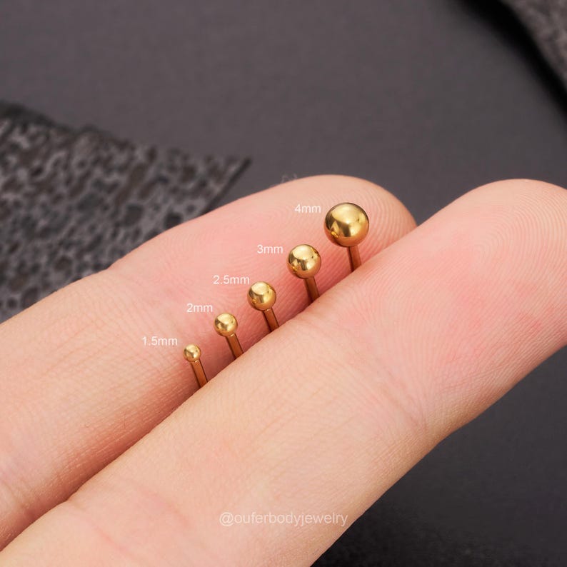 May include: A set of six gold-colored ball end studs in different sizes, ranging from 1.5mm to 4mm. The studs are arranged in order of size, with the smallest on the left and the largest on the right. The studs are held in a hand, with the background blurred.