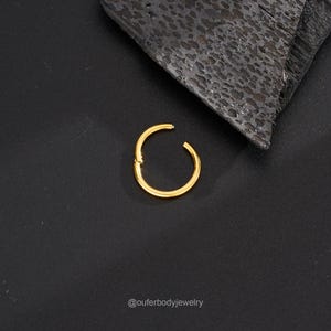 May include: Gold hinged seamless ring for body piercing. The ring is closed and lying on a black surface.