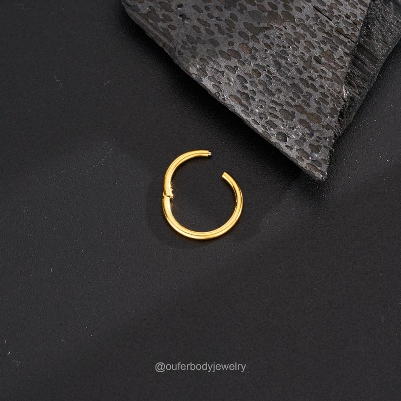 May include: Gold hinged seamless ring for body piercing. The ring is closed and lying on a black surface.