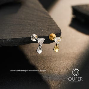 May include: Two labret piercings featuring a heart-shaped clear gemstone stud with a small dangling heart charm. One piece is silver-toned and the other is gold-toned. The text "Search OuferJewelry for more stunning designs!" and "OUFER BODY JEWELRY" appear.