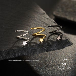 Peut inclure: Trois ear cuffs en spirale argentés, dorés et noirs, chacun avec un charme en forme d'étoile. Les ear cuffs sont présentés sur une surface sombre et texturée. Le texte "Search OuferJewelry for more stunning designs!" est visible.