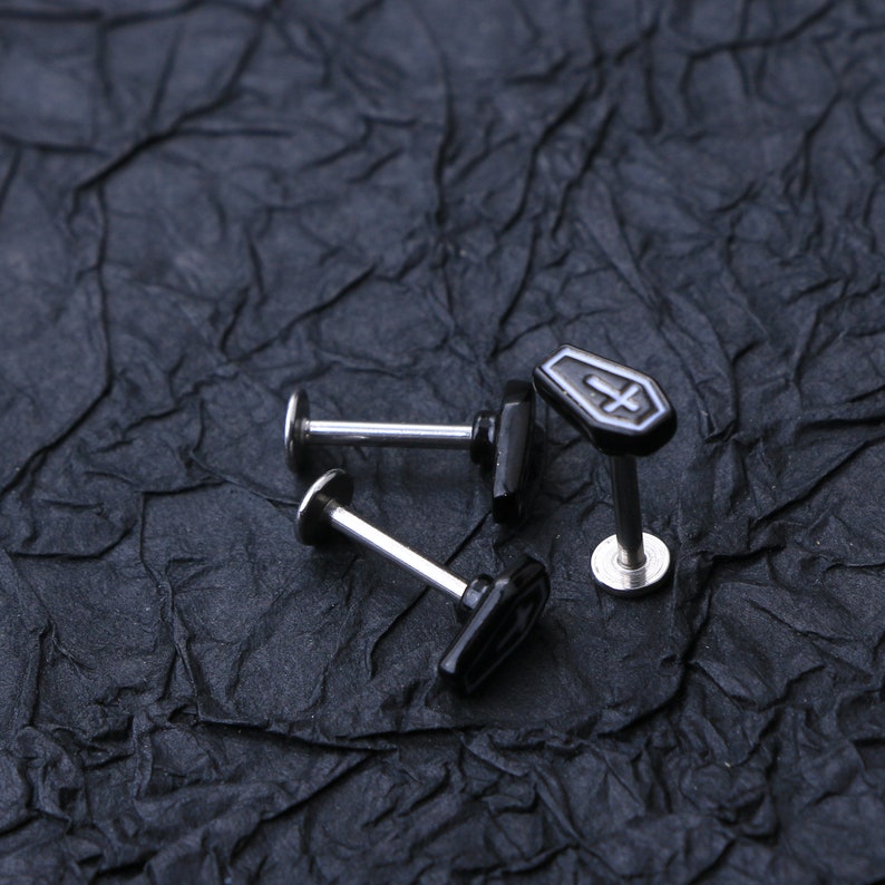 May include: Three silver barbell earrings with black coffin-shaped charms. The charms have a white cross design.