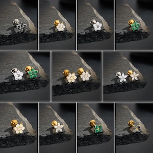 Puede incluir: Una colecci&oacute;n de pendientes de bot&oacute;n con forma de flor. Los pendientes presentan piedras preciosas de varios colores, incluyendo transparente, verde y negro. Los pendientes tienen postes plateados o dorados. Los pendientes se muestran sobre una superficie oscura y texturizada.