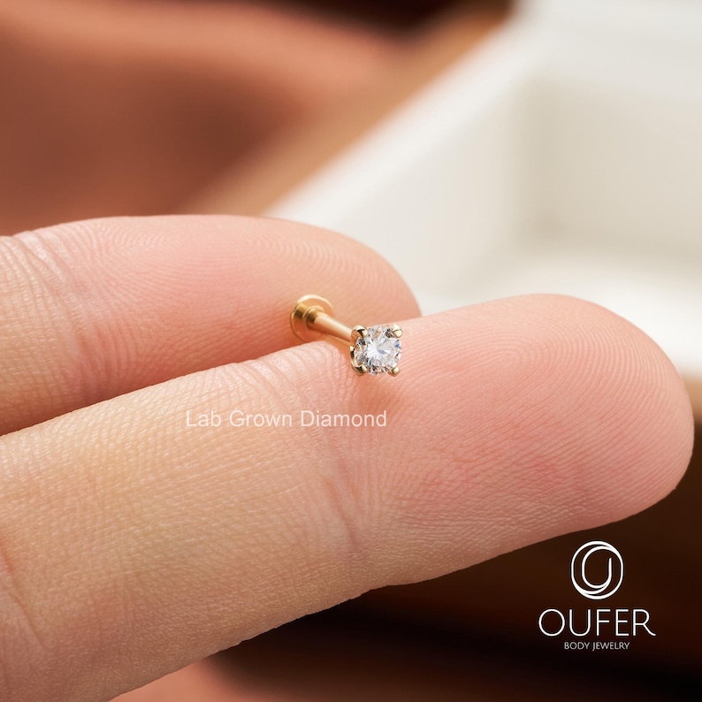 Puede incluir: Un pendiente de bot&oacute;n de diamante cultivado en laboratorio de color dorado. El pendiente presenta un peque&ntilde;o diamante de talla cuadrada engastado en un poste plano. El texto "Lab Grown Diamond" es visible. El pendiente se sostiene entre dos dedos.