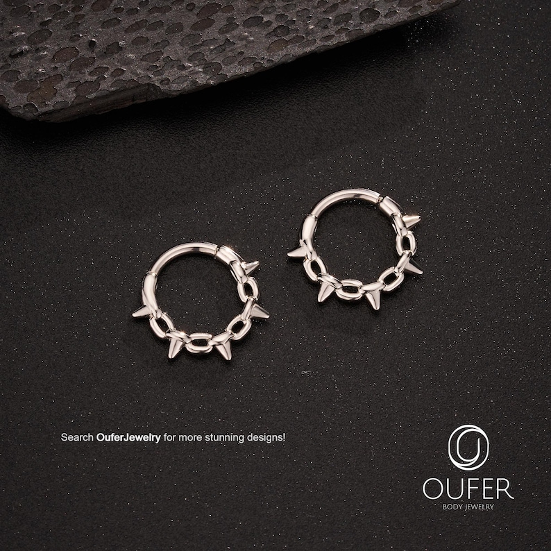 Op de afbeelding: Twee zilverkleurige oorringen met een ketting van kleine, puntige spikes. De oorbellen liggen op een zwart oppervlak. De tekst "Search OuferJewelry for more stunning designs!" staat onderaan de afbeelding.