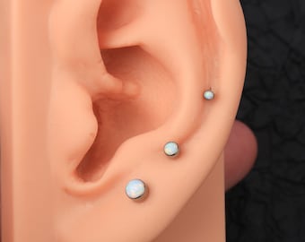 Puede incluir: Tres pendientes de ópalo en un piercing de cartílago. Los pendientes son de plata con una piedra de ópalo blanca.
