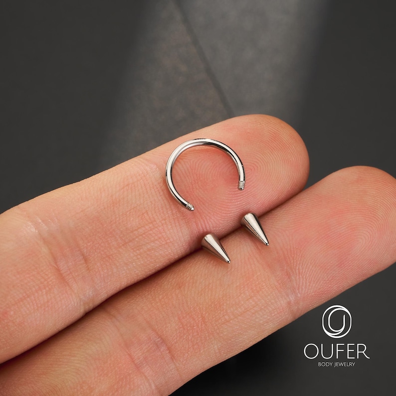 May include: A silver-colored circular horseshoe barbell body jewelry piece featuring two detachable cone-shaped ends with threaded posts. The metal ring has a smooth, polished finish and is designed for piercings, displaying the OUFER brand logo.