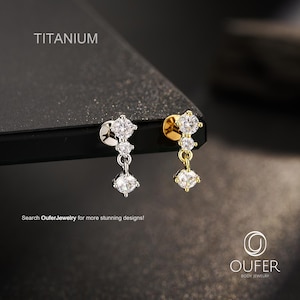 May include: Two titanium body jewelry studs with clear round-cut stones. One stud is silver-toned and the other is gold-toned. Each features a vertical arrangement of two stones with a third dangling stone below. Text reads TITANIUM and OUFER.