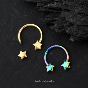 May include: Two gold and rainbow colored horseshoe shaped body jewelry pieces with star shaped ends. The jewelry is on a black background.