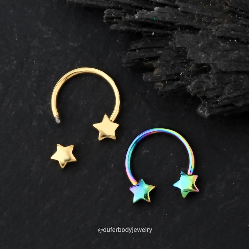 May include: Two gold and rainbow colored horseshoe shaped body jewelry pieces with star shaped ends. The jewelry is on a black background.