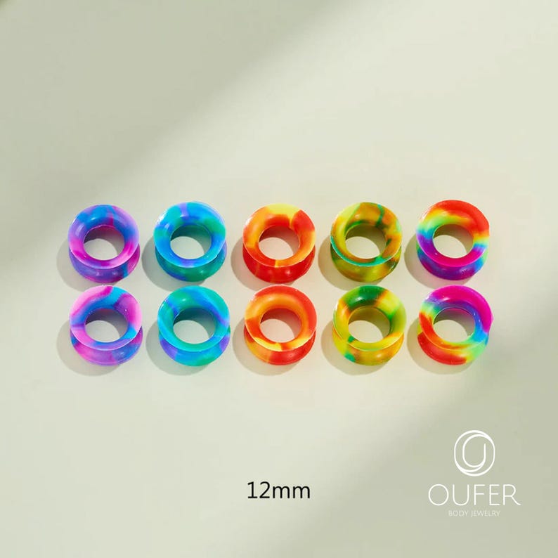 May include: Ten colorful, rainbow-colored ear gauges. Each gauge is a circular tunnel design, with a 12mm diameter. The gauges are in shades of blue, green, yellow, orange, and pink. The brand name "OUFER BODY JEWELRY" is in the bottom right corner.