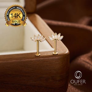 Pode incluir: Dois brincos labret de ouro 14K com um topo em forma de leque adornado com cinco pedras transparentes de corte marquise. As peças têm uma haste reta e um disco traseiro plano. Um selo dourado com 14K GOLD e a marca OUFER são visíveis.