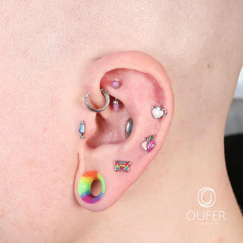 May include: An ear with multiple piercings, showcasing various types of body jewelry. The jewelry includes a large rainbow-colored ear plug, a silver hoop, and several small studs with colorful gems and designs. The ear is against a neutral background.