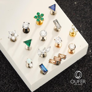 Puede incluir: Una colecci&oacute;n de pendientes de joyer&iacute;a corporal en varios dise&ntilde;os. Los pendientes presentan piedras preciosas en tonos claros, verdes, azules y &aacute;mbar, engastadas en metal plateado y dorado. Los dise&ntilde;os incluyen tri&aacute;ngulos, flores y formas geom&eacute;tricas.