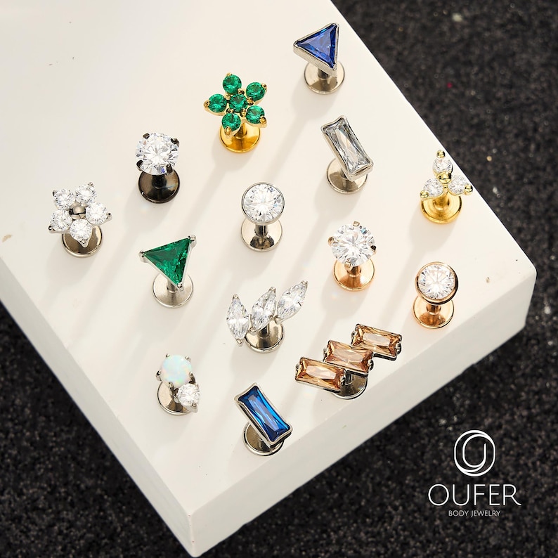 Puede incluir: Una colecci&oacute;n de pendientes de joyer&iacute;a corporal en varios dise&ntilde;os. Los pendientes presentan piedras preciosas en tonos claros, verdes, azules y &aacute;mbar, engastadas en metal plateado y dorado. Los dise&ntilde;os incluyen tri&aacute;ngulos, flores y formas geom&eacute;tricas.