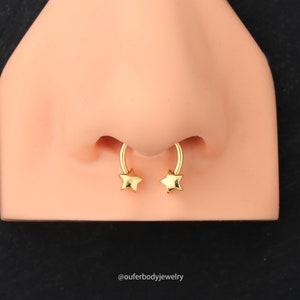 May include: Gold nose ring with star-shaped ends. The ring is curved and has a horseshoe shape.