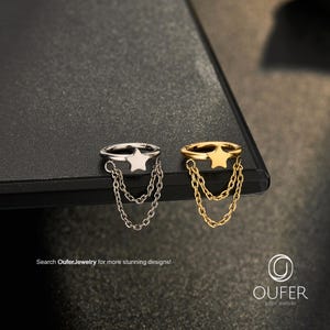 Peut inclure: Deux anneaux circulaires, l'un argenté et l'autre doré, chacun doté d'une petite breloque en forme d'étoile attachée à deux fines chaînes pendantes. Le texte "Search OuferJewelry for more stunning designs!" et le logo "OUFER BODY JEWELRY" sont visibles.