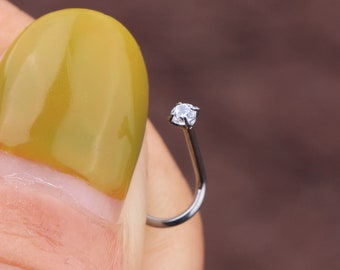May include: A silver nose ring featuring a curved post and a small, clear, round gemstone. The gemstone is set in a four-prong setting. The nose ring is held by a person with yellow nail polish.