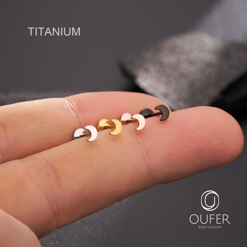 May include: A selection of crescent moon-shaped body jewellery in silver, gold, rose gold, and black finishes. The word "TITANIUM" is printed in white. The jewellery is displayed on a finger.
