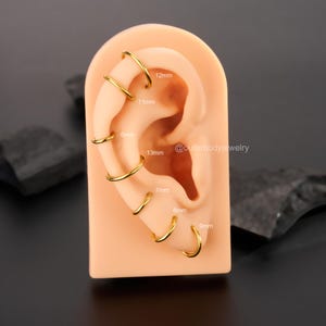 May include: A close-up of a model ear with multiple gold hoop earrings in different sizes. The earrings are labeled with their diameter in millimeters: 6mm, 7mm, 8mm, 9mm, 11mm, 12mm, and 13mm. The text "@outerbodyjewelry" is also visible on the model ear.