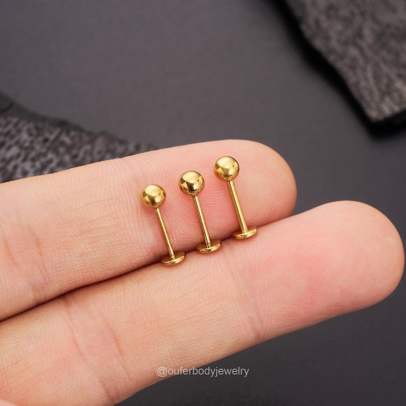 May include: Three gold-colored barbell studs with round ends. The studs are arranged in a row, with the largest stud on the left and the smallest stud on the right.