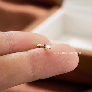 Puede incluir: Un peque&ntilde;o pendiente de bot&oacute;n de color dorado con una gema cuadrada y transparente. El pendiente se sostiene entre los dedos, con el texto "Lab Grown Diamond" visible en el fondo. Es una joya.