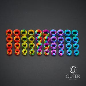 May include: A collection of colorful ear gauges in various sizes. The gauges display a rainbow of colors, including red, orange, yellow, green, blue, and purple. The body jewelry is arranged neatly on a dark surface.