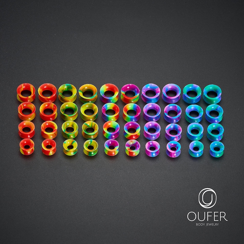 May include: A collection of colorful ear gauges in various sizes. The gauges display a rainbow of colors, including red, orange, yellow, green, blue, and purple. The body jewelry is arranged neatly on a dark surface.