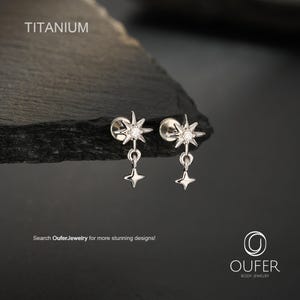 Puede incluir: Un par de pendientes de titanio en tono plateado con diseño de estrella, una piedra central transparente y un colgante de estrella de cuatro puntas. El texto TITANIUM es visible arriba, y el texto Search OuferJewelry for more stunning designs! y OUFER BODY JEWELRY están en la parte inferior.