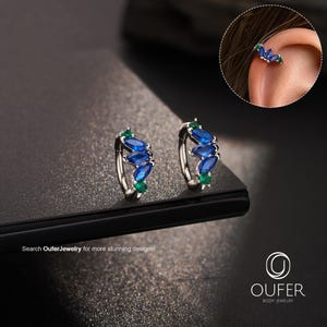 May include: A pair of silver-toned hoop earrings featuring marquise-cut blue stones and round green stones. The jewelry is branded with the text Oufer Body Jewelry and the phrase Search OuferJewelry for more stunning designs.