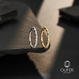 May include: Two circular hoop rings, one in a silver tone and one in a gold tone, featuring a twisted rope-like texture along the entire band. The rings are positioned upright, side by side, and include the text OUFER BODY JEWELRY in the corner.