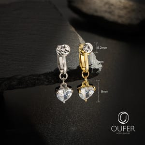 May include: Two clip-on belly rings, one silver-toned and one gold-toned, each featuring a clear heart-shaped crystal charm. The jewelry includes technical labels indicating a 5.2mm top diameter, 7.5mm length, and 9mm heart charm height.