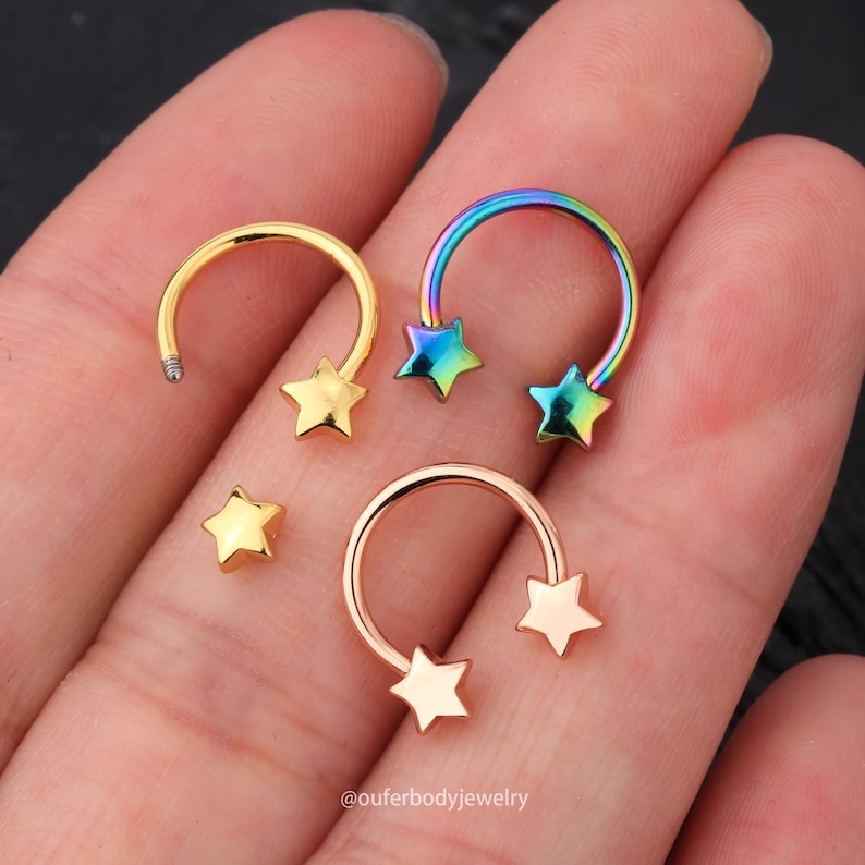 May include: Three gold and rainbow colored star-shaped horseshoe rings. The rings are for body piercings. The rings are on a hand.