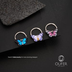 May include: Three silver body jewellery rings, each with a colourful butterfly design. The butterflies display shades of blue, purple, and pink. The text "Search OuferJewelry for more stunning designs!" is visible.