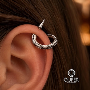 Op de afbeelding: Een zilverkleurige kraakbeenpiercing in de vorm van een gebogen octopus-tentakel met textuur van zuignappen. Het sieraad heeft een gladde, puntige sluiting aan de bovenkant. Het logo OUFER BODY JEWELRY is zichtbaar in de rechterbenedenhoek.