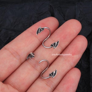 Peut inclure: Deux boucles d'oreilles helix en argent avec des motifs d'ailes de chauve-souris noires. Les boucles d'oreilles sont pr&eacute;sent&eacute;es sur une main.