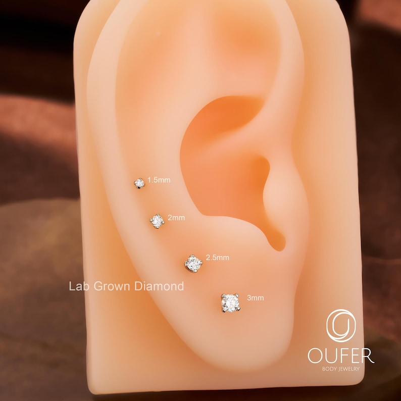 Puede incluir: Una exhibici&oacute;n de pendientes de diamantes cultivados en laboratorio en varios tama&ntilde;os, de 1,5 mm a 3 mm, presentados en un modelo de oreja realista. Los pendientes son de corte redondo y brillantes, mostrando el dise&ntilde;o de la joyer&iacute;a. El logotipo de la marca es visible.