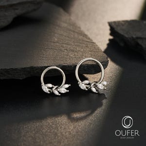 Può includere: Due orecchini a cerchio color argento con un gruppo di pietre trasparenti taglio marquise disposte a forma di foglia lungo il bordo inferiore. Il testo OUFER BODY JEWELRY con un logo circolare è presente nell'angolo in basso a destra.