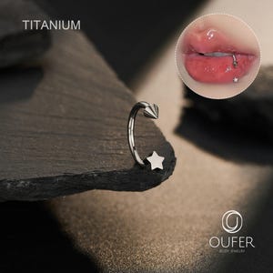 May include: A silver-colored body piercing jewelry with a star and cone design. The word "TITANIUM" is printed at the top. The jewelry is displayed on a dark, textured surface. A close-up of a lip piercing is shown in the upper right corner.
