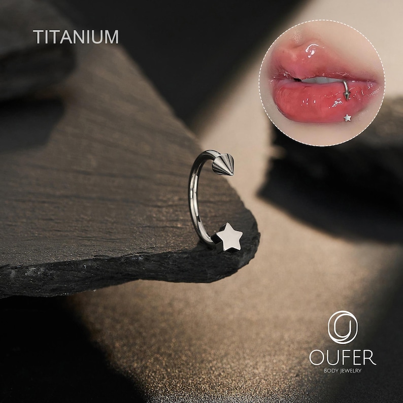 May include: A silver-colored body piercing jewelry with a star and cone design. The word "TITANIUM" is printed at the top. The jewelry is displayed on a dark, textured surface. A close-up of a lip piercing is shown in the upper right corner.
