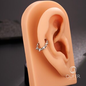 Op de afbeelding: Een zilveren oorring met kleine, puntige spikes wordt getoond op een huidkleurig oormodel. De oorring is een doorlopende lus en de spikes zijn gelijkmatig verdeeld over de omtrek. De achtergrond is donkergrijs.