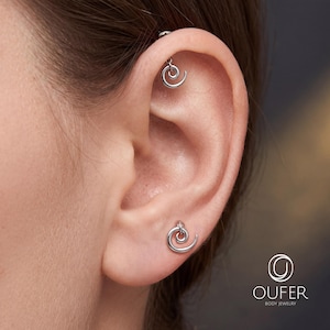 Può includere: Un paio di orecchini color argento a forma di spirale. Un orecchino è posizionato nella cartilagine superiore dell'orecchio, mentre l'altro si trova sul lobo. Il logo OUFER BODY JEWELRY è visibile nell'angolo in basso a destra dell'immagine.