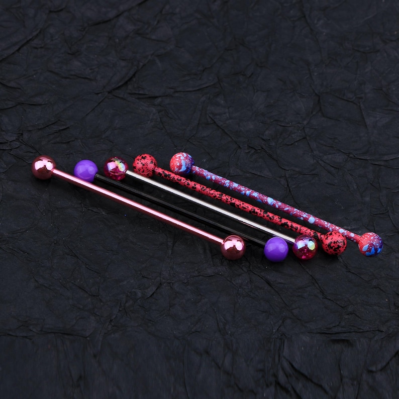 May include: Four different barbell piercings with colorful balls. The barbells are made of metal and have a variety of colors, including pink, purple, and black. The balls are also colorful and have a variety of designs.